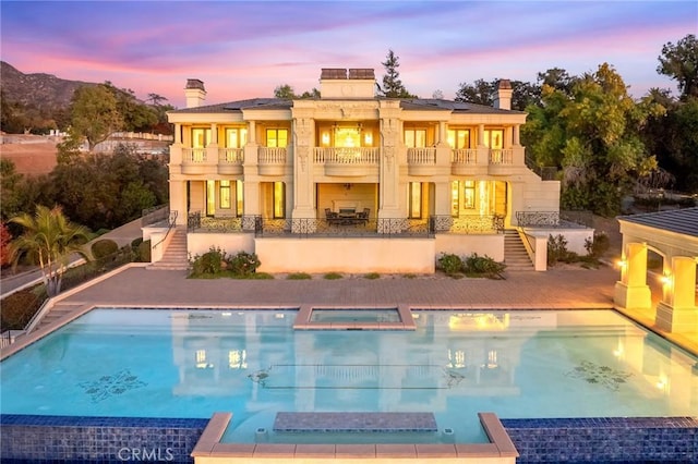 back house at dusk with a swimming pool with hot tub, a balcony, and a patio