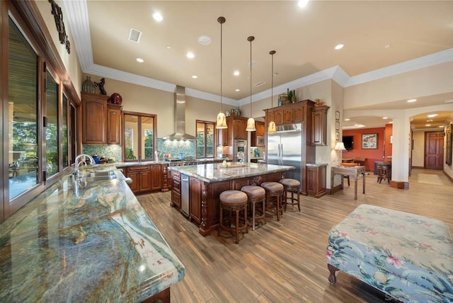 kitchen with sink, wall chimney range hood, built in refrigerator, a spacious island, and decorative light fixtures