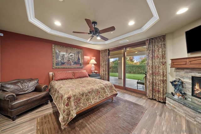 bedroom with access to exterior, ceiling fan, a stone fireplace, light hardwood / wood-style floors, and a tray ceiling