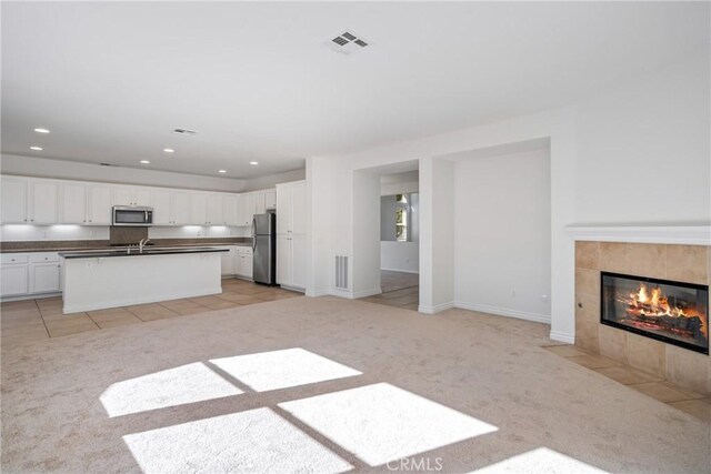 kitchen with white cabinetry, stainless steel appliances, light carpet, a tiled fireplace, and a kitchen island with sink