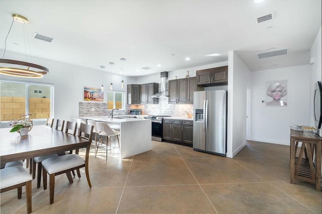 kitchen with a breakfast bar, wall chimney range hood, hanging light fixtures, dark brown cabinets, and stainless steel appliances