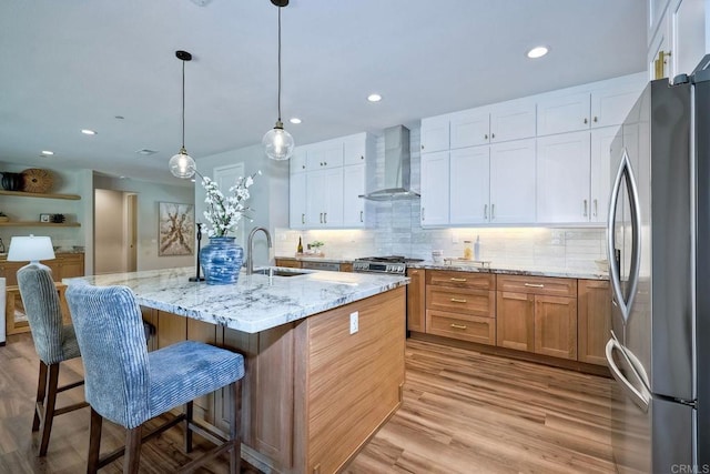 kitchen with stainless steel appliances, sink, wall chimney range hood, white cabinets, and an island with sink