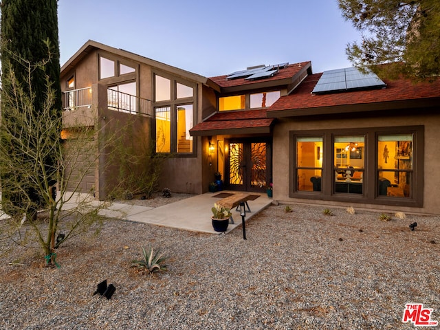 back house at dusk featuring solar panels, a balcony, and a patio