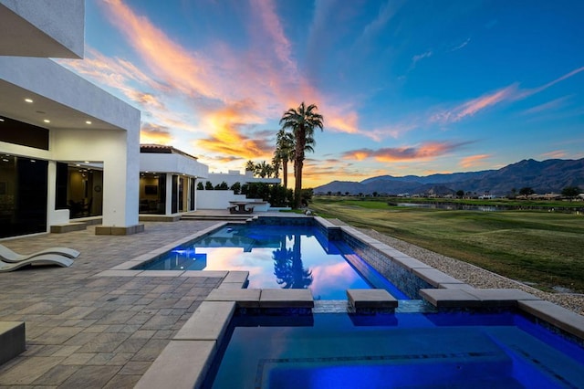 pool at dusk with an in ground hot tub, a mountain view, a yard, and a patio area