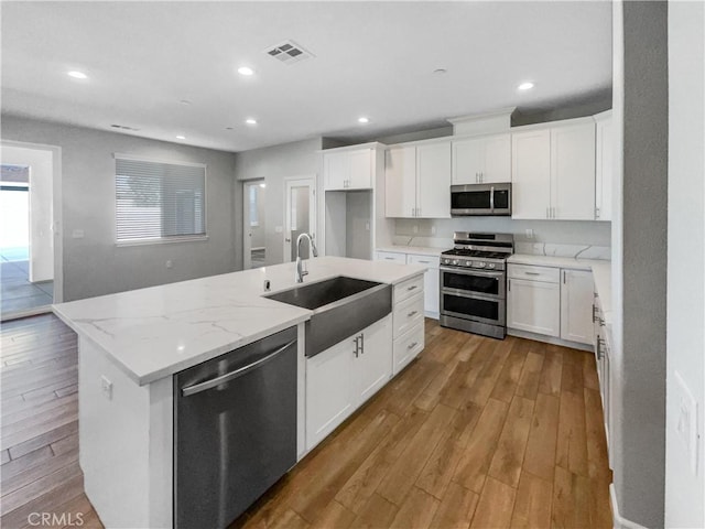 kitchen with sink, appliances with stainless steel finishes, a kitchen island with sink, light stone counters, and white cabinets