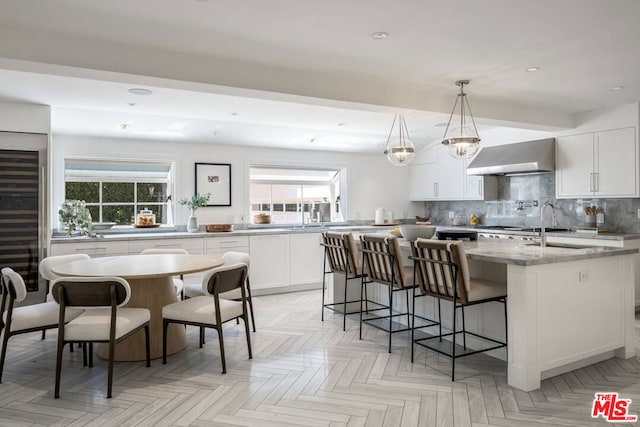 kitchen featuring white cabinets, decorative backsplash, an island with sink, range hood, and decorative light fixtures