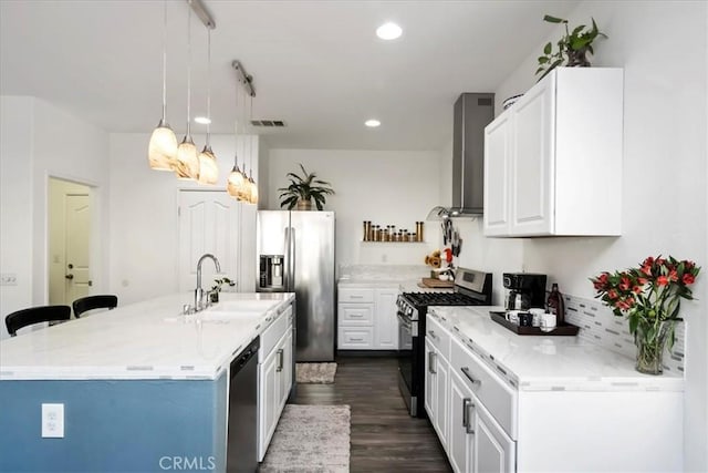 kitchen featuring white cabinets, wall chimney range hood, hanging light fixtures, an island with sink, and stainless steel appliances