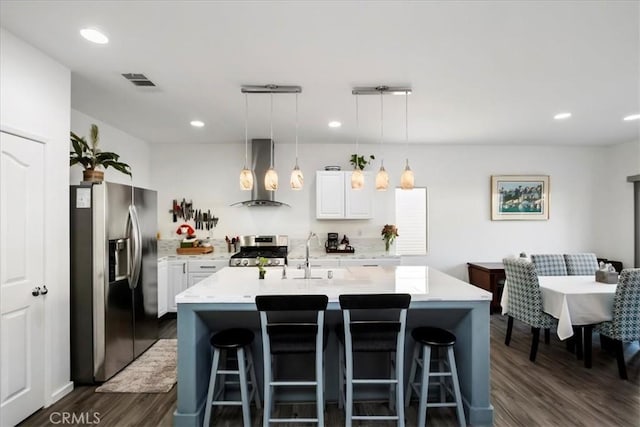 kitchen featuring stainless steel appliances, wall chimney range hood, white cabinetry, hanging light fixtures, and an island with sink