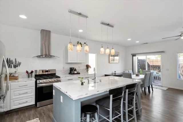 kitchen featuring hanging light fixtures, stainless steel gas range, wall chimney exhaust hood, an island with sink, and white cabinetry