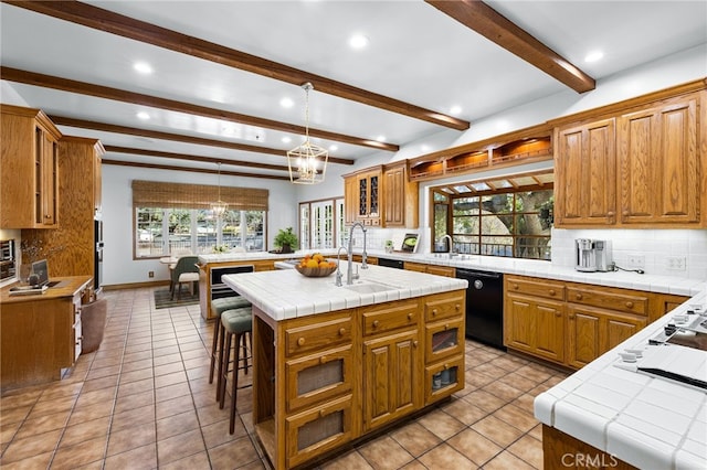 kitchen with dishwasher, tile counters, sink, a kitchen island with sink, and beam ceiling