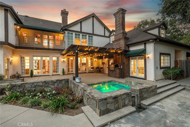 back house at dusk with a pergola, a patio area, exterior kitchen, a balcony, and an in ground hot tub