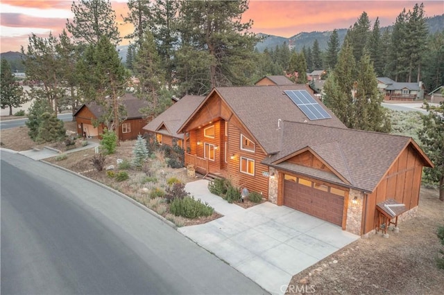 view of front facade with a garage, driveway, stone siding, roof with shingles, and roof mounted solar panels