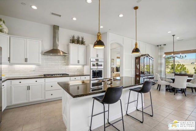 kitchen featuring white cabinets, stainless steel appliances, a kitchen island with sink, and wall chimney range hood