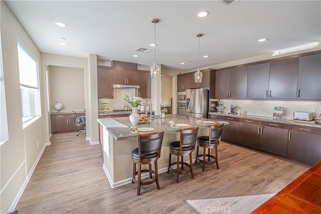 kitchen with dark brown cabinetry, light hardwood / wood-style flooring, backsplash, an island with sink, and appliances with stainless steel finishes