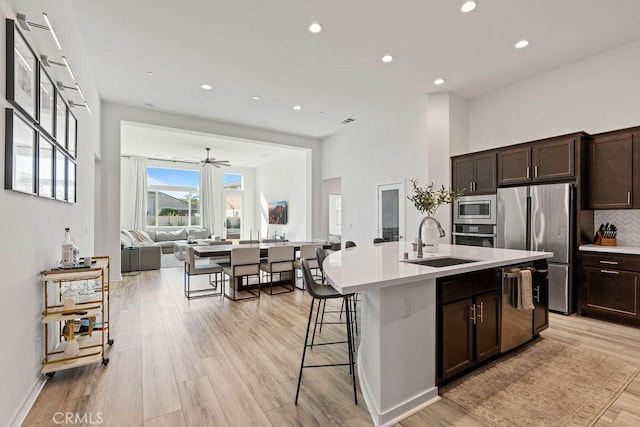 kitchen with sink, light wood-type flooring, an island with sink, dark brown cabinets, and stainless steel appliances