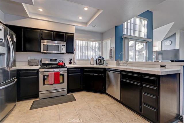 kitchen featuring tile countertops, a raised ceiling, light tile patterned floors, kitchen peninsula, and stainless steel appliances