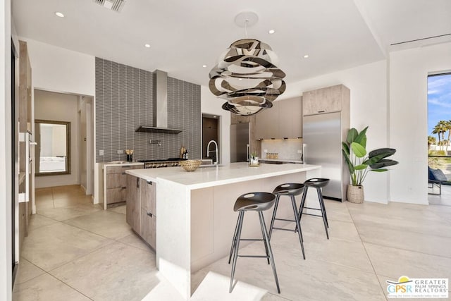 kitchen with tasteful backsplash, built in refrigerator, a kitchen island with sink, and wall chimney range hood