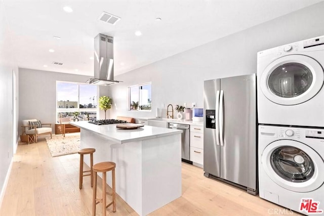 kitchen featuring light hardwood / wood-style flooring, appliances with stainless steel finishes, stacked washer / drying machine, island range hood, and white cabinetry