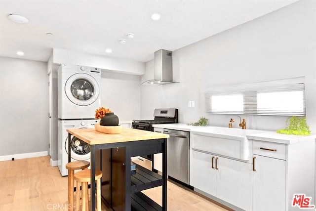 kitchen featuring light wood-type flooring, wall chimney exhaust hood, stainless steel appliances, white cabinetry, and stacked washer / drying machine