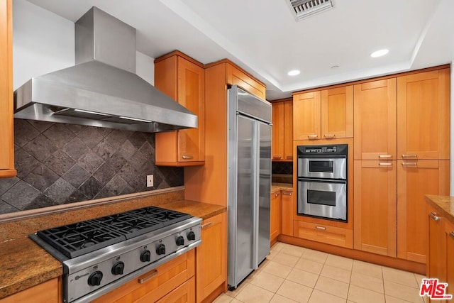 kitchen with wall chimney exhaust hood, light tile patterned floors, dark stone counters, stainless steel appliances, and backsplash