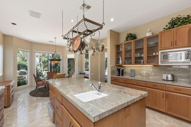 kitchen featuring light stone countertops, stainless steel microwave, sink, decorative backsplash, and a center island with sink