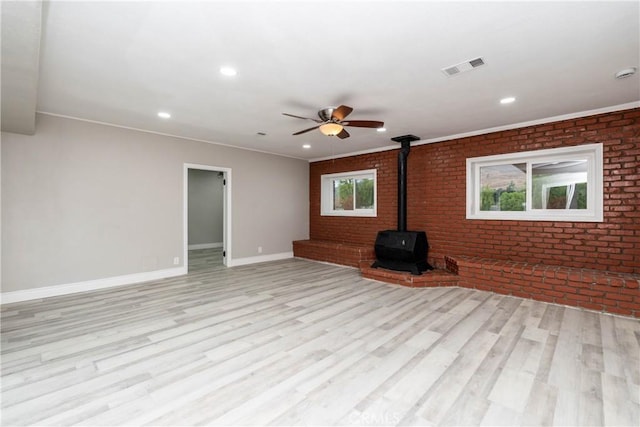 unfurnished living room with a wood stove, ceiling fan, brick wall, and light wood-type flooring