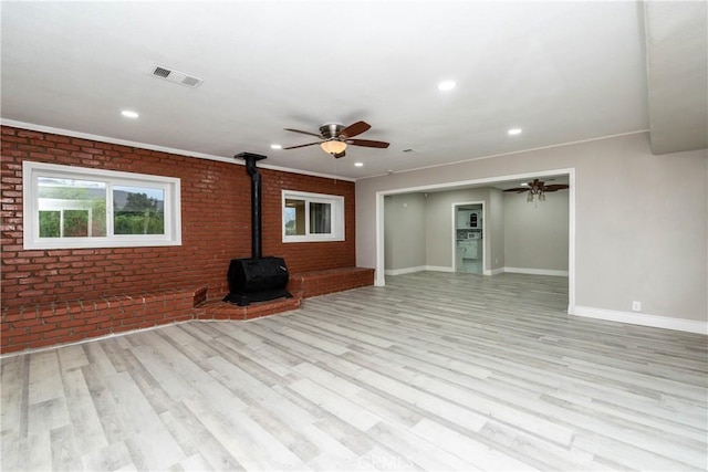 unfurnished living room with a wood stove, ceiling fan, brick wall, light wood-type flooring, and ornamental molding