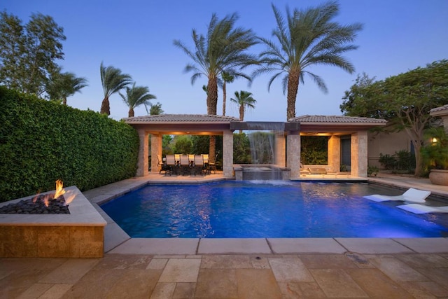 pool at dusk featuring an in ground hot tub, pool water feature, a fire pit, and a patio area