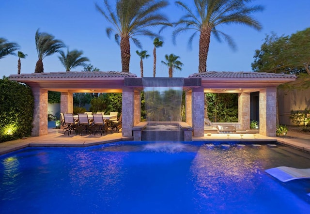 pool at dusk featuring pool water feature, a patio, and a hot tub