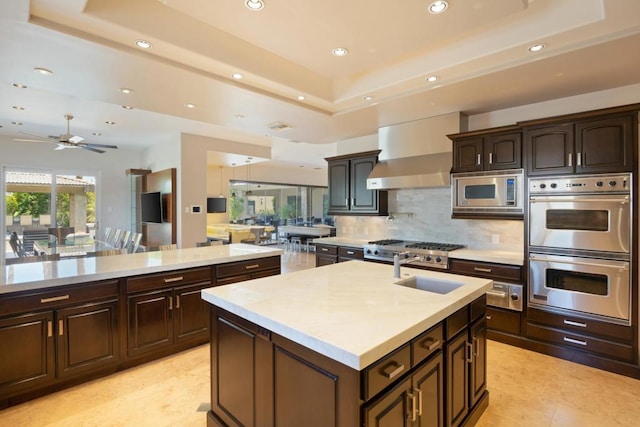 kitchen with ceiling fan, tasteful backsplash, a raised ceiling, an island with sink, and appliances with stainless steel finishes