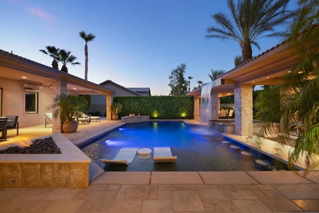 pool at dusk featuring a patio area, pool water feature, and an outdoor fire pit