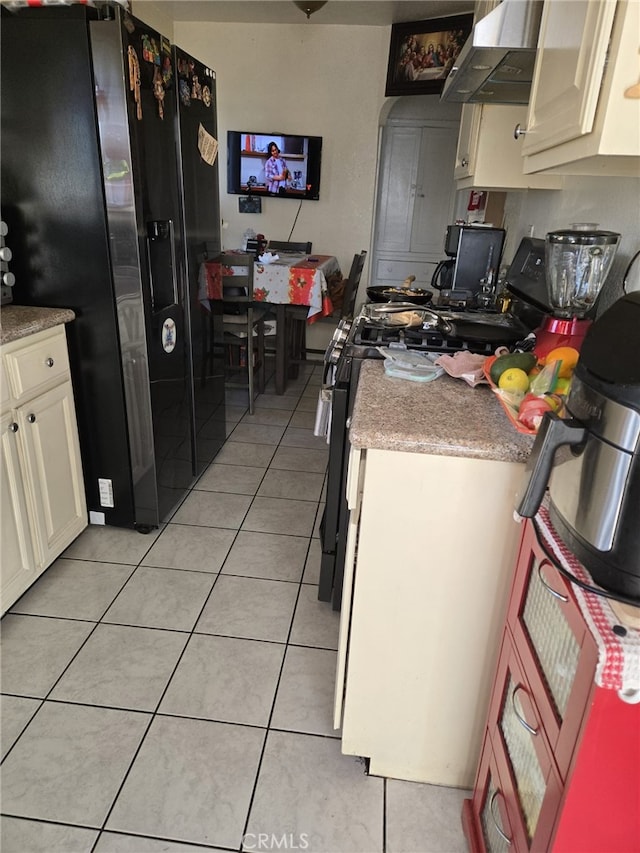 kitchen with range, cream cabinetry, light tile patterned floors, black fridge, and wall chimney range hood