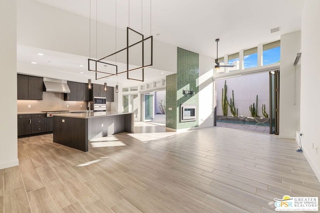 kitchen with a wealth of natural light, dark brown cabinets, a kitchen island with sink, and wall chimney range hood