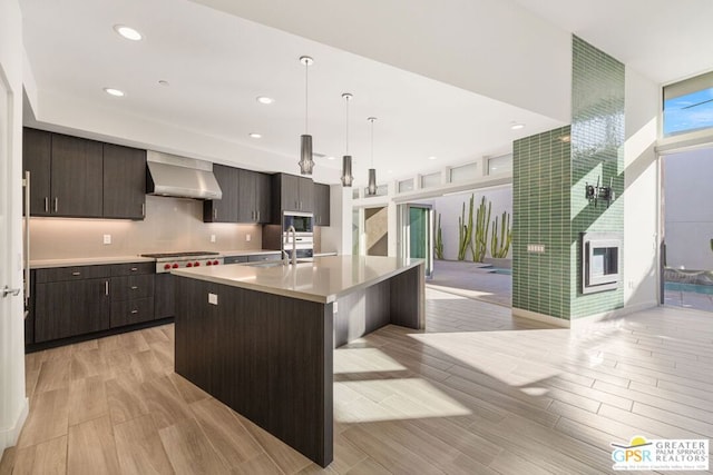 kitchen with stainless steel appliances, hanging light fixtures, wall chimney range hood, a kitchen island with sink, and light wood-type flooring