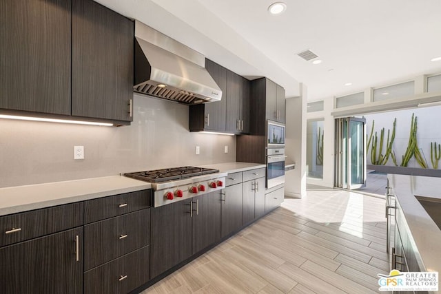 kitchen featuring dark brown cabinetry, light hardwood / wood-style flooring, wall chimney range hood, and appliances with stainless steel finishes