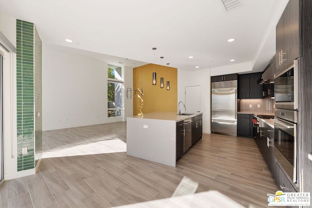 kitchen with a center island with sink, sink, light hardwood / wood-style flooring, wall chimney exhaust hood, and appliances with stainless steel finishes