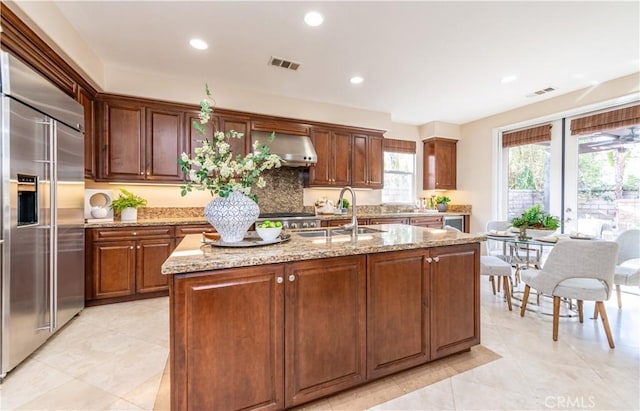 kitchen featuring a kitchen island with sink, sink, built in refrigerator, wall chimney exhaust hood, and light stone countertops