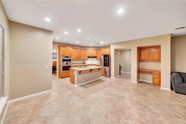 kitchen with appliances with stainless steel finishes, backsplash, built in desk, a center island with sink, and a breakfast bar area