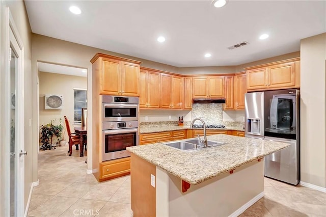 kitchen with light stone counters, stainless steel appliances, sink, light tile patterned floors, and a center island with sink