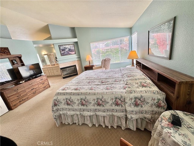 bedroom featuring light carpet, a glass covered fireplace, connected bathroom, lofted ceiling, and a textured wall