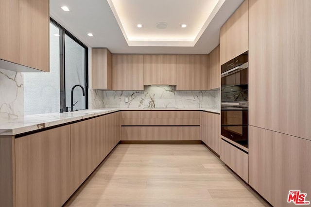 kitchen featuring light brown cabinets, backsplash, a raised ceiling, light hardwood / wood-style flooring, and light stone counters