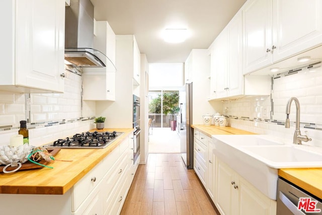 kitchen featuring appliances with stainless steel finishes, white cabinetry, wall chimney range hood, butcher block counters, and sink