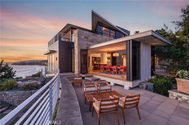 back house at dusk with a balcony, a water view, and a patio area