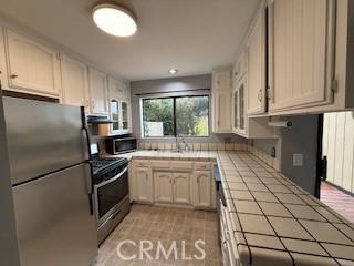 kitchen with tile counters, sink, stainless steel appliances, white cabinets, and exhaust hood