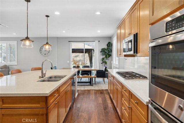 kitchen with dark wood-type flooring, hanging light fixtures, a kitchen island with sink, appliances with stainless steel finishes, and sink