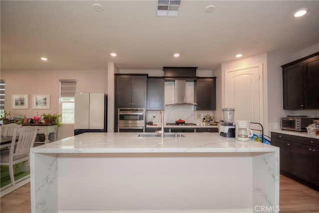 kitchen with appliances with stainless steel finishes, a kitchen island with sink, sink, wall chimney range hood, and light hardwood / wood-style flooring