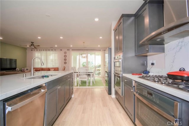 kitchen featuring sink, hanging light fixtures, wall chimney range hood, light hardwood / wood-style flooring, and appliances with stainless steel finishes