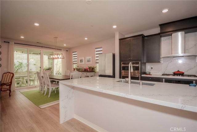 kitchen with stainless steel appliances, wall chimney range hood, light stone counters, pendant lighting, and light hardwood / wood-style floors