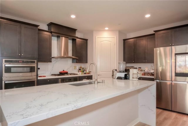 kitchen with appliances with stainless steel finishes, wall chimney exhaust hood, a kitchen island with sink, sink, and light hardwood / wood-style flooring