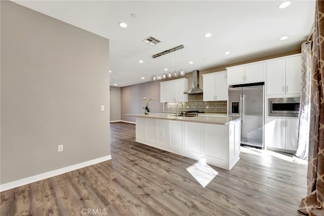 kitchen with appliances with stainless steel finishes, wall chimney range hood, a center island with sink, decorative light fixtures, and light hardwood / wood-style floors